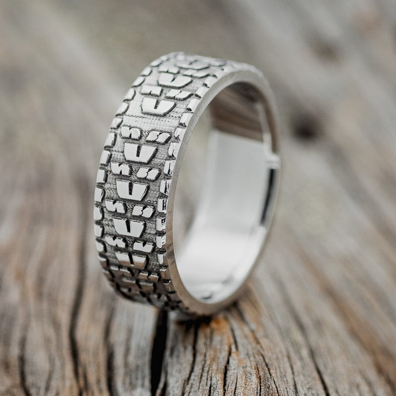 Shown here is "Parcel", a handcrafted, custom embossed men's wedding ring featuring a dirt bike tire tread engraving, upright facing left. This ring is pictured in a titanium band. It can be customized to feature just about any embossed design you can dream up.