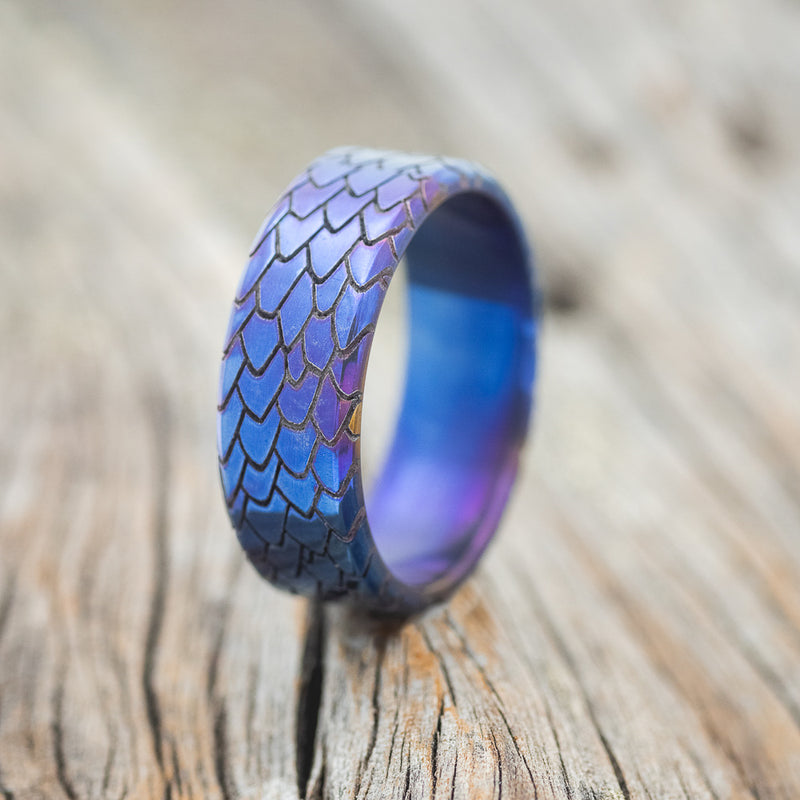 Shown here is "Echo", a handcrafted, custom embossed titanium men's wedding ring featuring a dragon scale engraving with a fire-treated finish, upright facing left. It can be customized to feature just about any embossed design you can dream up.