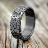 Shown here is "Echo", a handcrafted, custom embossed men's wedding ring featuring a dragon scale engraving, upright facing left. This ring is pictured in black zirconium, giving it a higher contrast between embossment and engraving than other metal types will provide. It can be customized to feature just about any embossed design you can dream up.