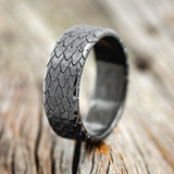 Shown here is "Echo", a handcrafted, custom embossed men's wedding ring featuring a dragon scale engraving with a hammered finish, upright facing left. This ring is pictured in black zirconium, giving it a higher contrast between embossment and engraving than other metal types will provide. It can be customized to feature just about any embossed design you can dream up.