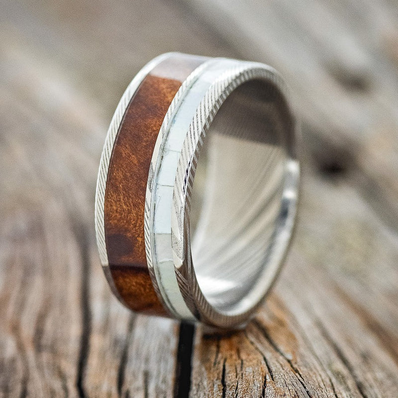 Shown here is "Raptor", a custom, handcrafted men's wedding ring featuring a mother of pearl & redwood inlay on a Damascus steel band, upright facing left. Additional inlay options are available upon request.