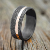 Shown here is "Vertigo", a handcrafted men's wedding ring featuring an offset wide 14K rose gold inlay and a hammered finish, upright facing left. Shown here set on a fire-treated black zirconium wedding band. Additional inlay options are available upon request.