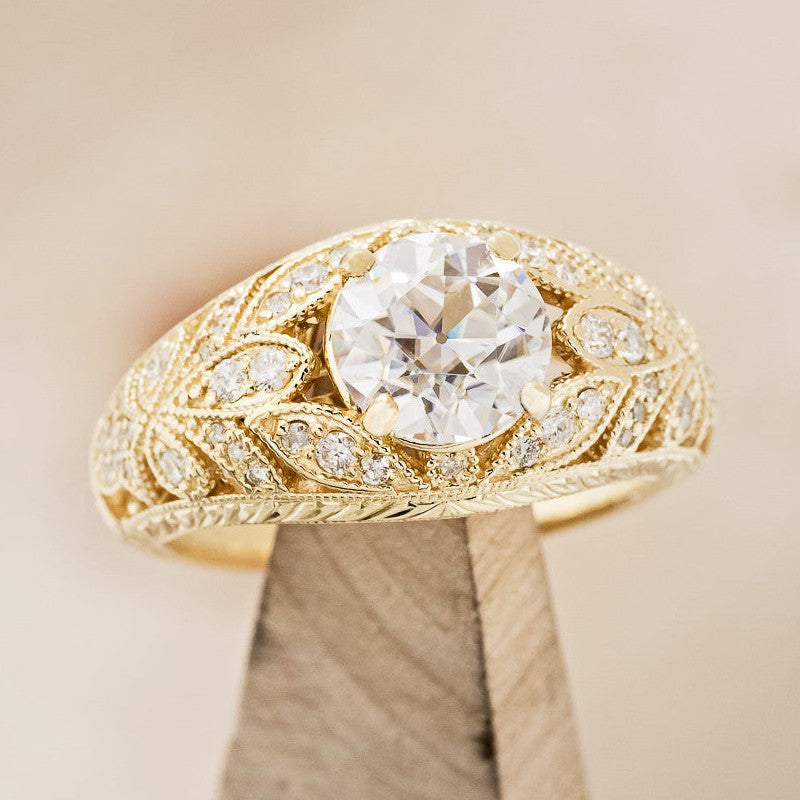 Shown here is The "Queen Of The Throne", an art-deco-style round moissanite women's engagement ring with delicate and ornate details and is available with many center stone options