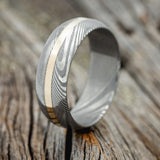 Shown here is "Nirvana", a custom handcrafted men's wedding ring featuring a domed profile Damascus steel band with a 14K yellow gold inlay, upright facing left. Additional inlay options are available upon request.