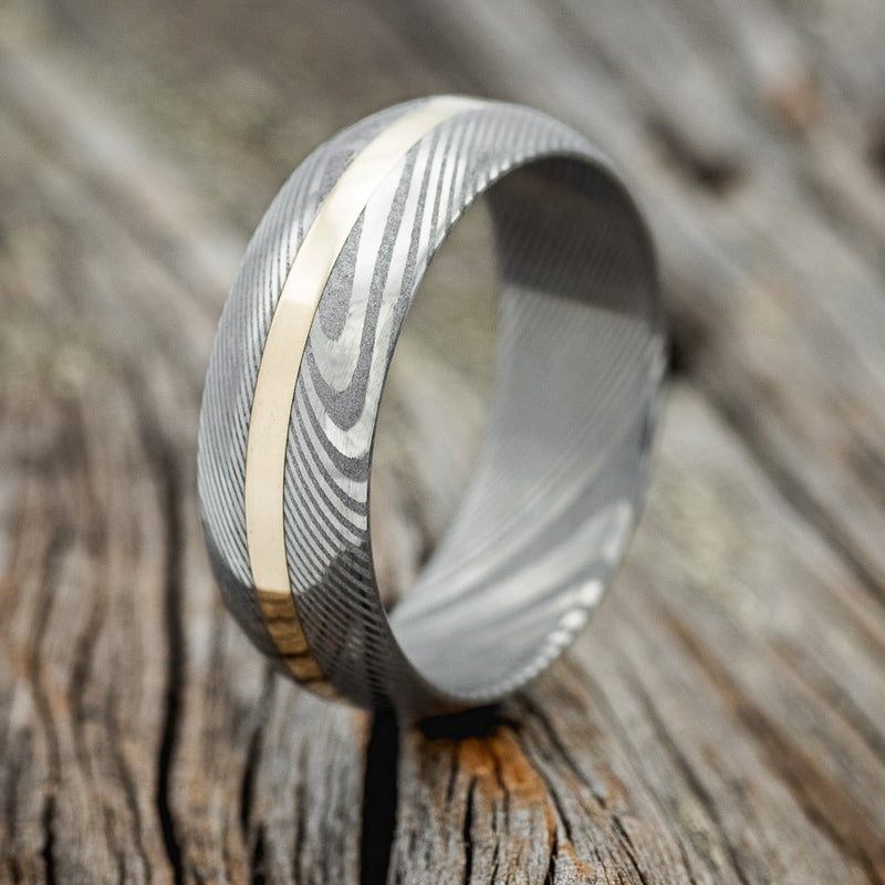 Shown here is "Nirvana", a custom handcrafted men's wedding ring featuring a domed profile Damascus steel band with a 14K yellow gold inlay, upright facing left. Additional inlay options are available upon request.