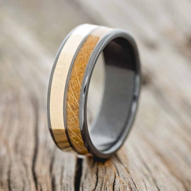 Shown here is "Dyad", a custom, handcrafted men's wedding ring featuring 2 channels with whiskey barrel oak and 14K yellow gold inlay, shown here on a fire-treated black zirconium band, upright facing left. Additional inlay options are available upon request.