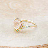 Shown here is "Dream", a rose quartz women's engagement ring with a diamond halo and diamond accents, facing left. Many other center stone options are available upon request.