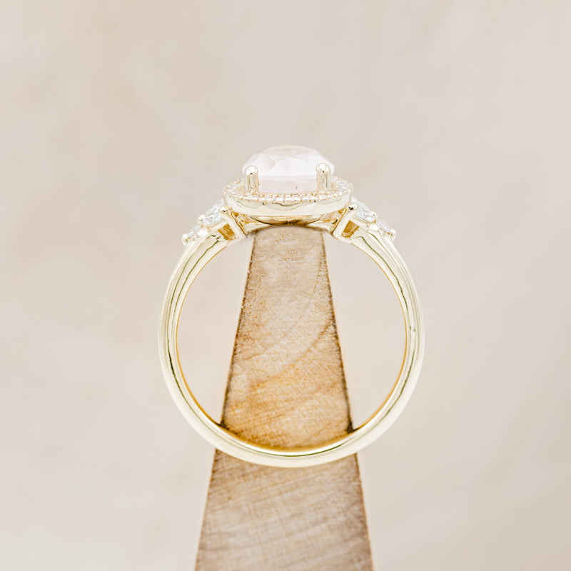 Shown here is "Dream", a rose quartz women's engagement ring with a diamond halo and diamond accents, side view on stand. Many other center stone options are available upon request.