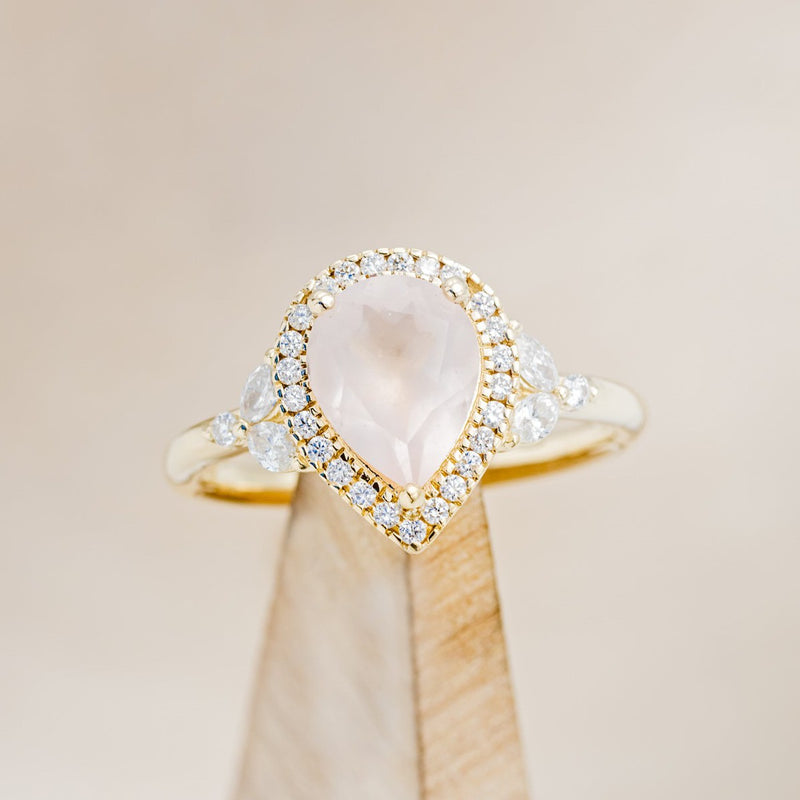 Shown here is "Dream", a rose quartz women's engagement ring with a diamond halo and diamond accents, on stand front facing. Many other center stone options are available upon request.