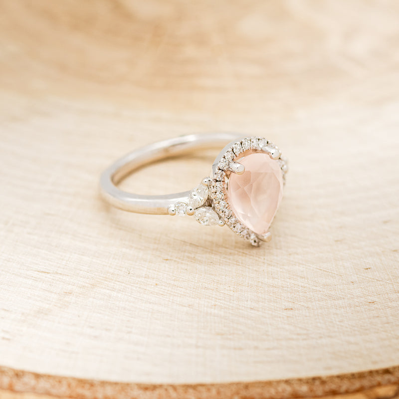 Shown here is "Dream", a rose quartz women's engagement ring with a diamond halo and diamond accents, facing right. Many other center stone options are available upon request.