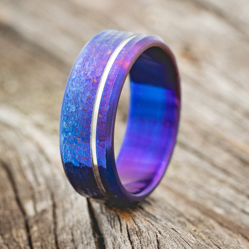 Shown here is a handcrafted men's wedding ring featuring a fire-treated titanium band with a cut etching and a hammered finish, upright facing left. 