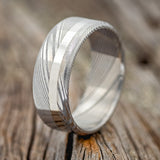 Shown here is "Vertigo", a custom handcrafted men's wedding ring featuring a Damascus steel band with a 14K white gold inlay, upright facing left. Additional inlay options are available upon request.