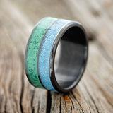 Shown here is "Dyad", a custom, handcrafted men's wedding ring featuring 2 channels with malachite and turquoise inlay, shown here set on a fire-treated black zirconium band, upright facing left. Additional inlay options are available upon request.