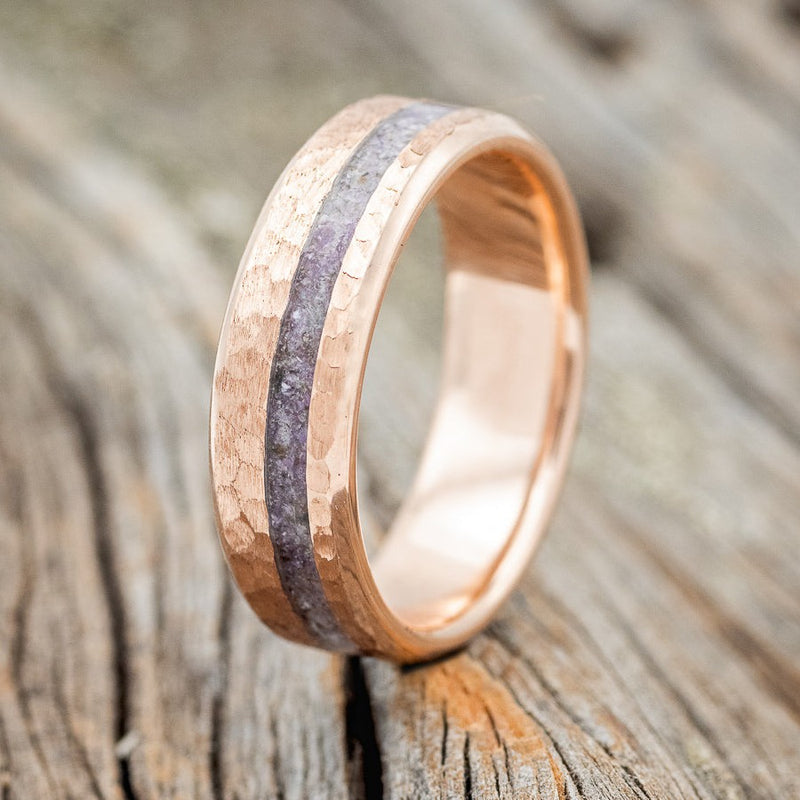 Shown here is "Vertigo", a custom, handcrafted men's wedding ring featuring a 14K gold wedding band with a charoite inlay and hammered finish, upright facing left. Additional inlay options are available upon request.