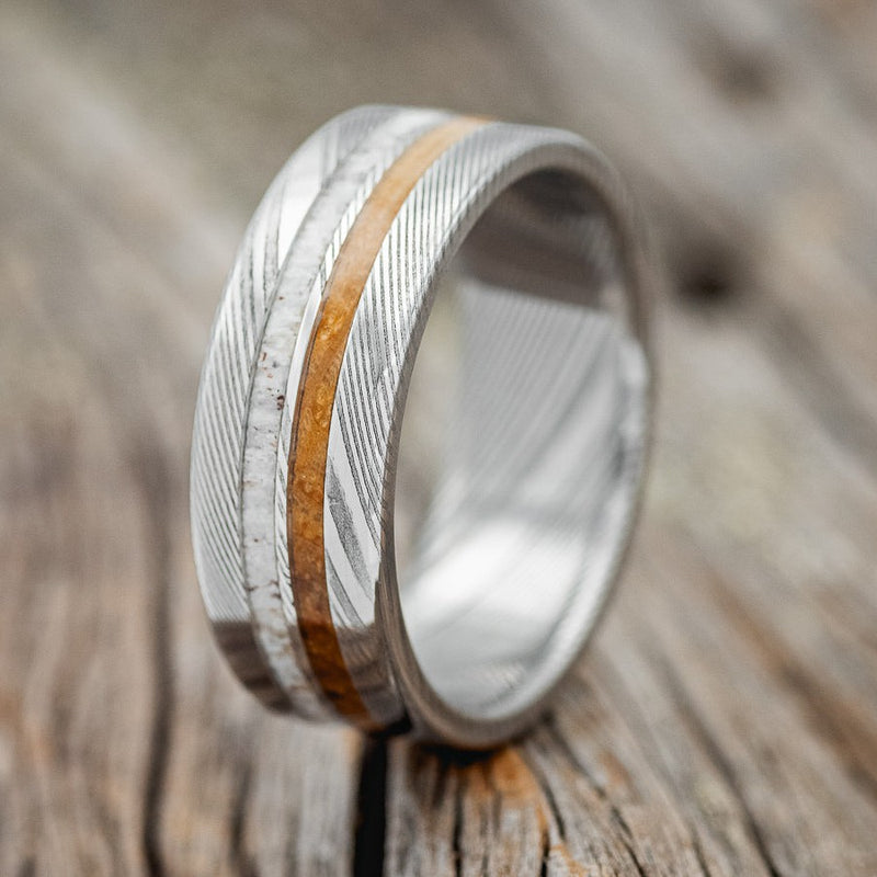 Shown here is "Silas", a custom, handcrafted men's wedding ring featuring whiskey barrel and antler inlays, shown here on a Damascus steel band, upright facing left. Additional inlay options are available upon request.