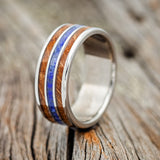 Shown here is "Rio", a custom, handcrafted men's wedding ring featuring 3 channels with a whiskey barrel and lapis lazuli inlays on a titanium band, upright facing left. Additional inlay options are available upon request.