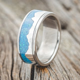 Shown here is "Helios", a custom, handcrafted men's wedding ring featuring a mountain range using pieces of silver and a blue opal inlay, upright facing left. Additional inlay options are available upon request.
