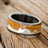 Shown here is "Helios", a custom, handcrafted men's wedding ring featuring a mountain range using pieces of silver and a whiskey barrel oak inlay, tilted left Additional inlay options are available upon request.