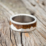 Shown here is "Helios", a custom, handcrafted men's wedding ring featuring a mountain range using pieces of silver and a whiskey barrel oak inlay, laying flat. Additional inlay options are available upon request.