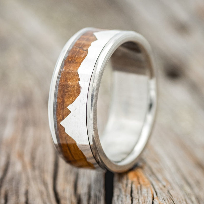 Shown here is "Helios", a custom, handcrafted men's wedding ring featuring a mountain range using pieces of silver and a whiskey barrel oak inlay, upright facing left. Additional inlay options are available upon request.