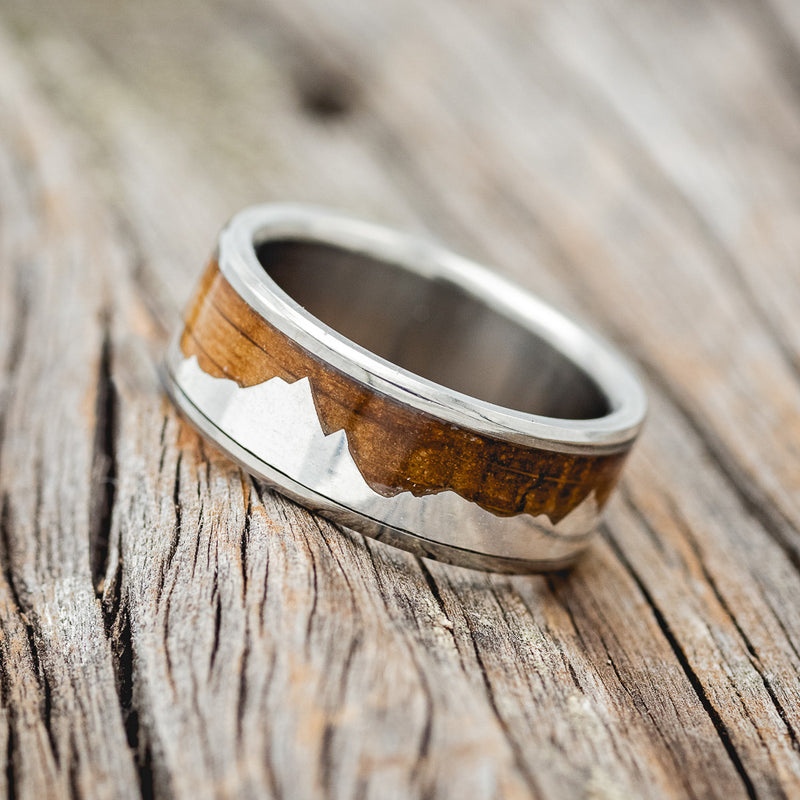 Shown here is "Helios", a custom, handcrafted men's wedding ring featuring a mountain range using pieces of silver and a whiskey barrel oak inlay, tilted left Additional inlay options are available upon request.