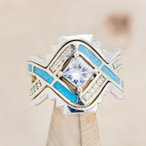 Shown here is "Helix", a geometric-style princess cut moissanite women's engagement ring with diamond accents and turquoise inlays, with tracers on stand front facing. Many other center stone options are available upon request.