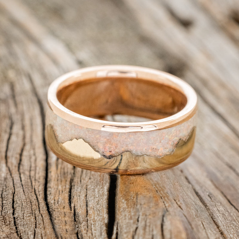 Shown here is "Helios", a custom, handcrafted men's wedding ring featuring a mountain range using pieces of gold and crushed fire & ice opal, laying flat. Additional inlay options are available upon request.