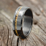 Shown here is "Vertigo", a custom, handcrafted men's wedding ring featuring a gold nugget inlay, shown here on a fire-treated black zirconium band, upright facing left. Additional inlay options are available upon request.