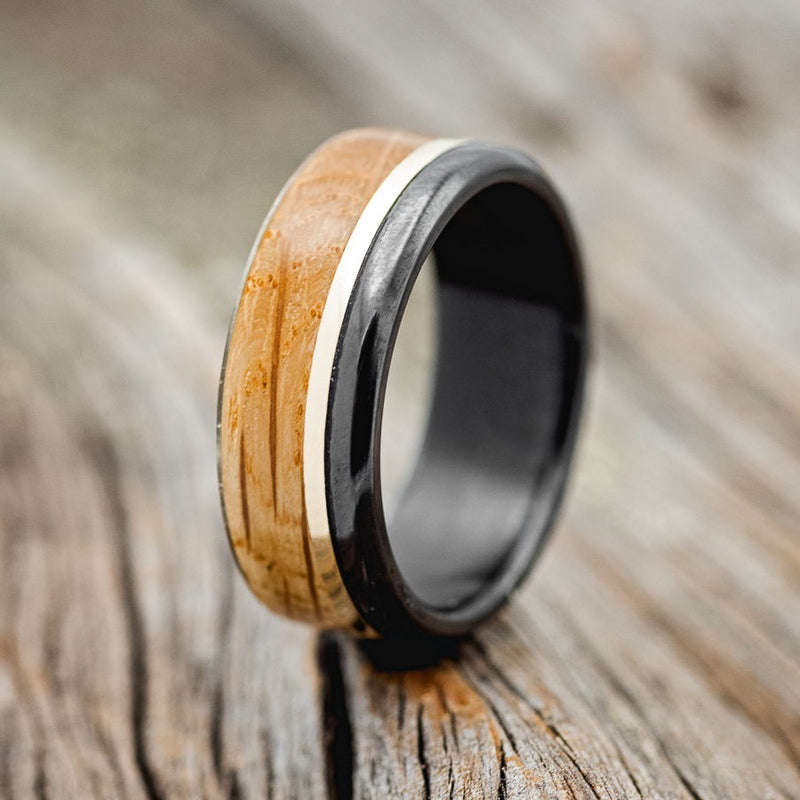 Shown here is "Ezra", a custom, handcrafted men's wedding ring featuring a whiskey barrel oak overlay and a 14K yellow gold inlay, shown here on a fire-treated black zirconium band, upright facing left. Additional inlay options are available upon request.