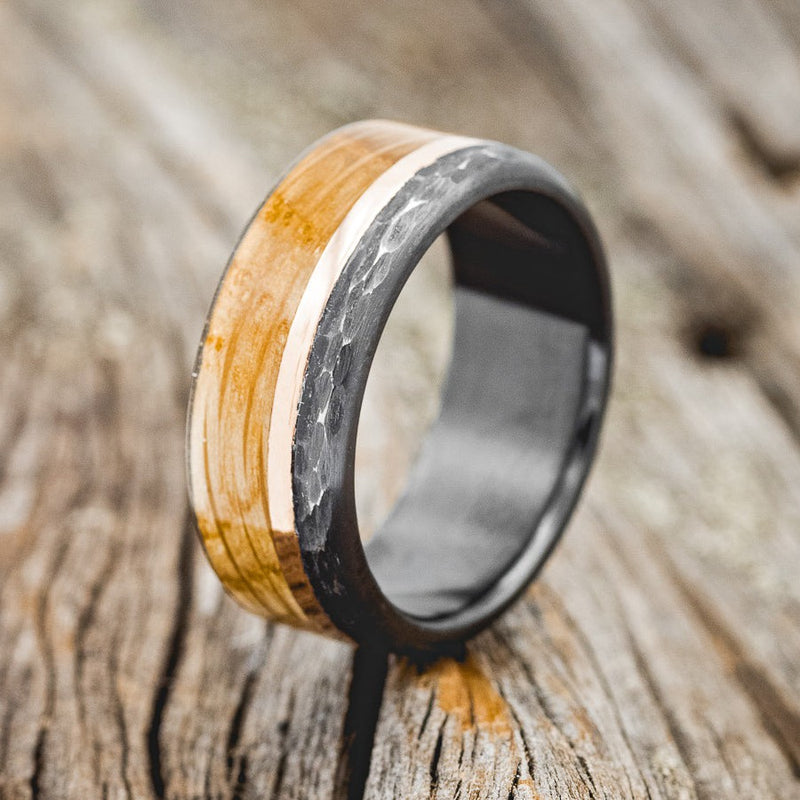Shown here is "Ezra", a custom, handcrafted men's wedding ring featuring a whiskey barrel oak overlay and a 14K rose gold inlay, shown here on a black zirconium band with a hammered finish, upright facing left. Additional inlay options are available upon request.
