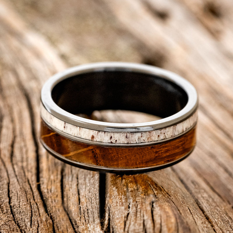 Shown here is "Raptor", a custom, handcrafted men's wedding ring featuring authentic whiskey barrel oak and elk antler on a fire-treated black zirconium band, laying flat. Additional inlay options are available upon request.