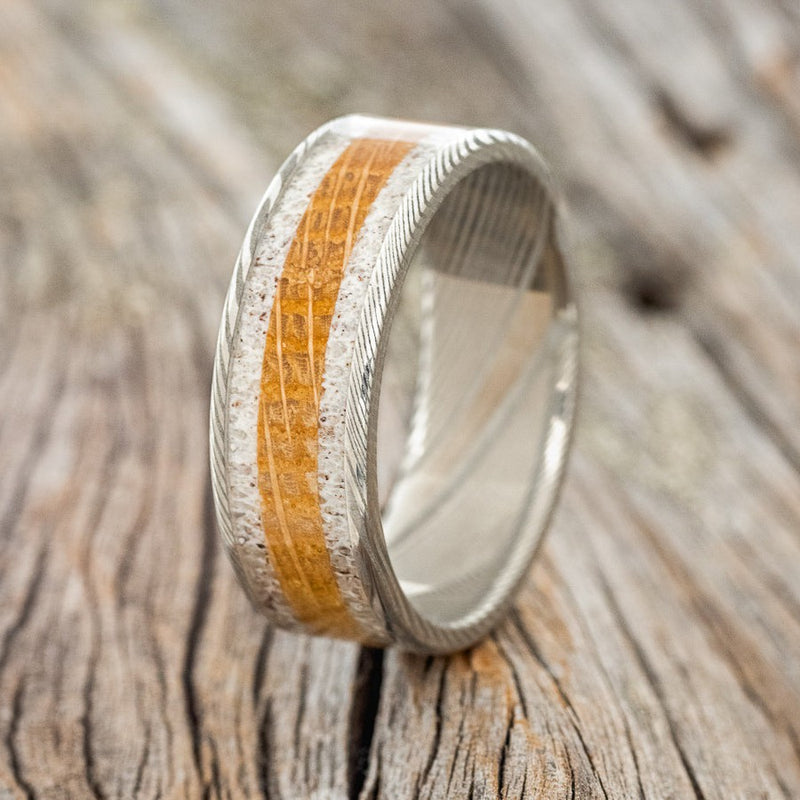 Shown here is "Rainier", a handcrafted men's wedding ring, shown featuring a Damascus Steel base with whiskey barrel and antler inlays, upright facing left. Additional inlay options are available upon request.