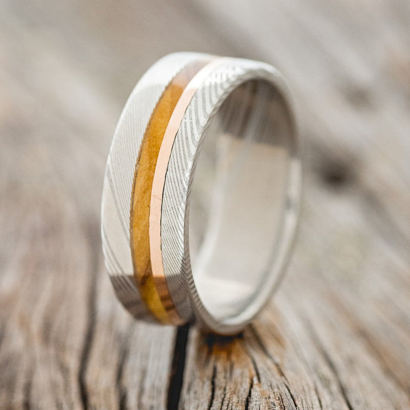 Shown here is a custom, handcrafted men's wedding ring featuring whiskey barrel oak and a 14K rose gold inlay, shown here on a Damascus steel band, upright facing left. Additional inlay options are available upon request.