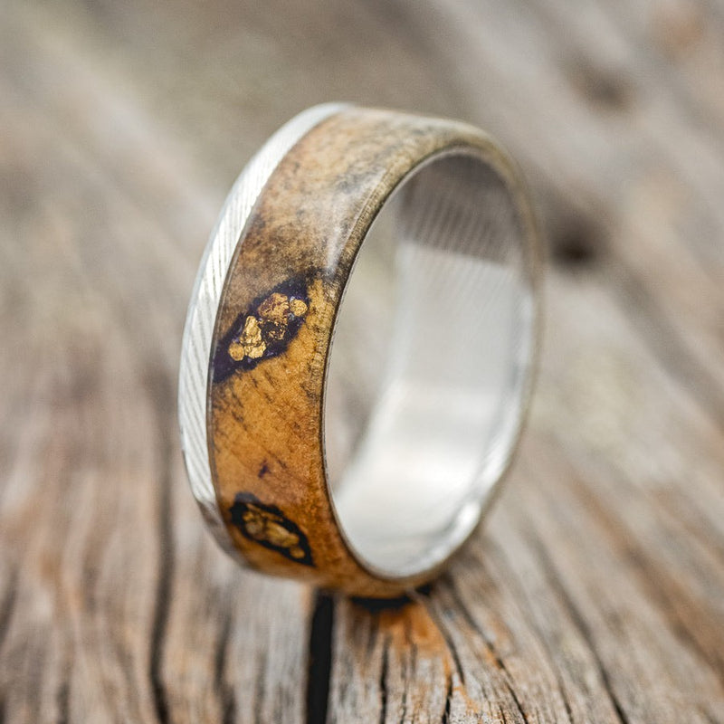 Shown here is "Ezra", a custom, handcrafted men's wedding ring featuring a buckeye burl wood overlay on a Damascus steel band with gold nuggets set in the burls, upright facing left. Additional inlay options are available upon request.