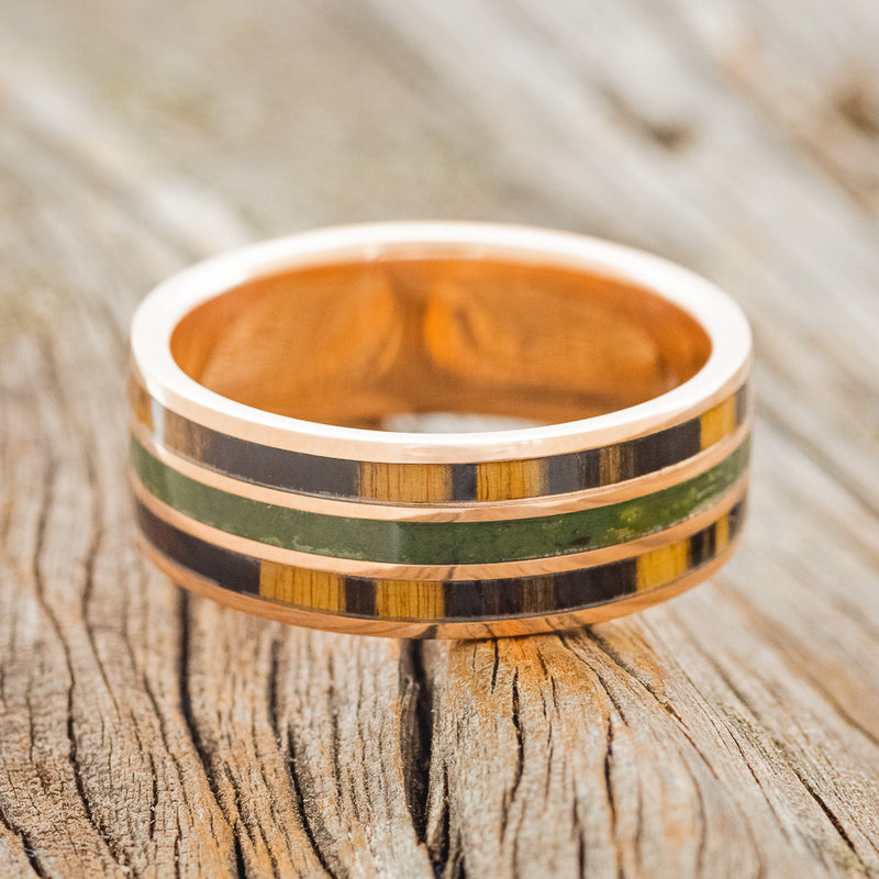 Shown here is "Rio", a custom, handcrafted men's wedding ring featuring 3 channels with white and black ebony wood and moss inlays on a 14K rose gold band, laying flat. Additional inlay options are available upon request.