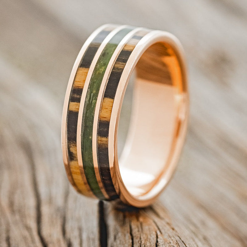 Shown here is "Rio", a custom, handcrafted men's wedding ring featuring 3 channels with white and black ebony wood and moss inlays on a 14K rose gold band, upright facing left. Additional inlay options are available upon request.