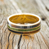 Shown here is "Rio", a custom, handcrafted men's wedding ring featuring 3 channels with white and black ebony wood and moss inlays on a 14K yellow gold band, laying flat. Additional inlay options are available upon request.