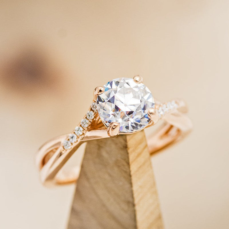 Shown here is "Roslyn", an accented-style round moissanite women's engagement ring, on stand facing slightly right, with delicate and ornate details and is available with many center stone options