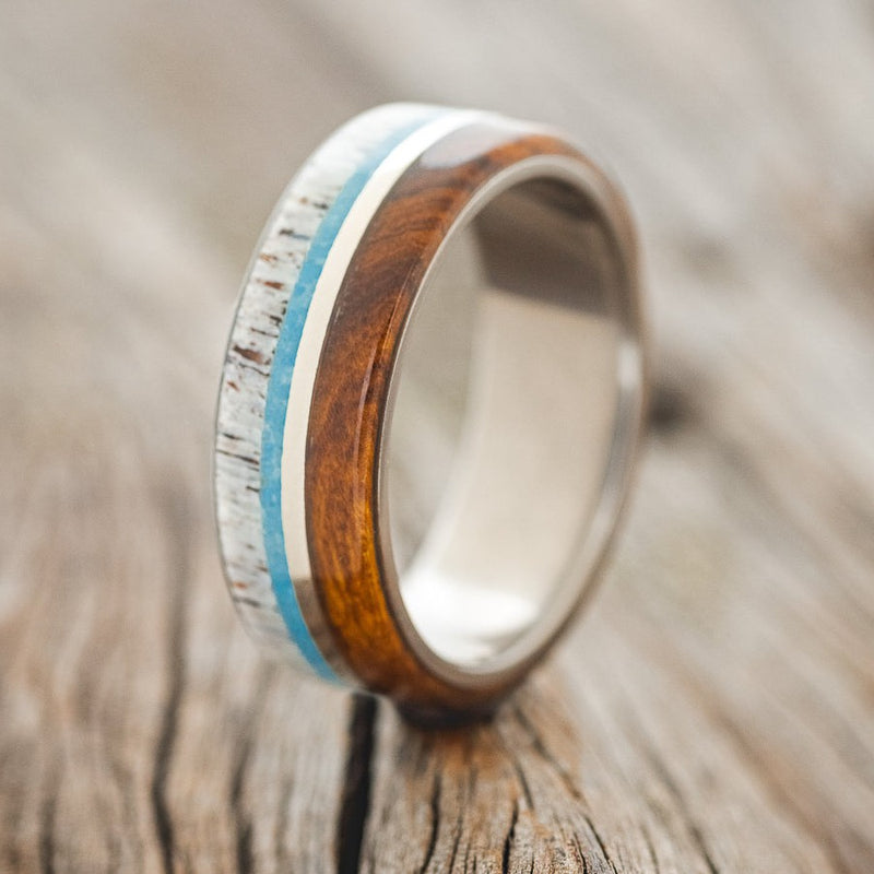 Shown here is "Banner", a custom, handcrafted men's wedding ring featuring naturally shed antler, turquoise, 14K white gold, and ironwood inlays shown here on a titanium band, upright facing left. Additional inlay options are available upon request.
