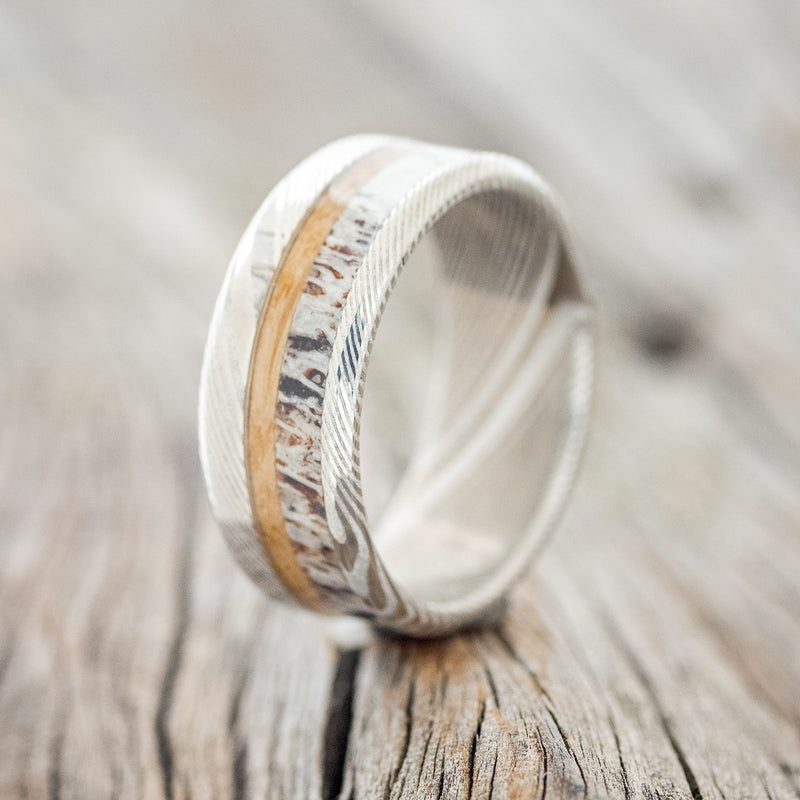 Shown here is "Tanner", a custom, handcrafted men's wedding ring featuring a whiskey barrel and antler inlay on a Damascus Steel band, upright facing left. Additional inlay options are available upon request.