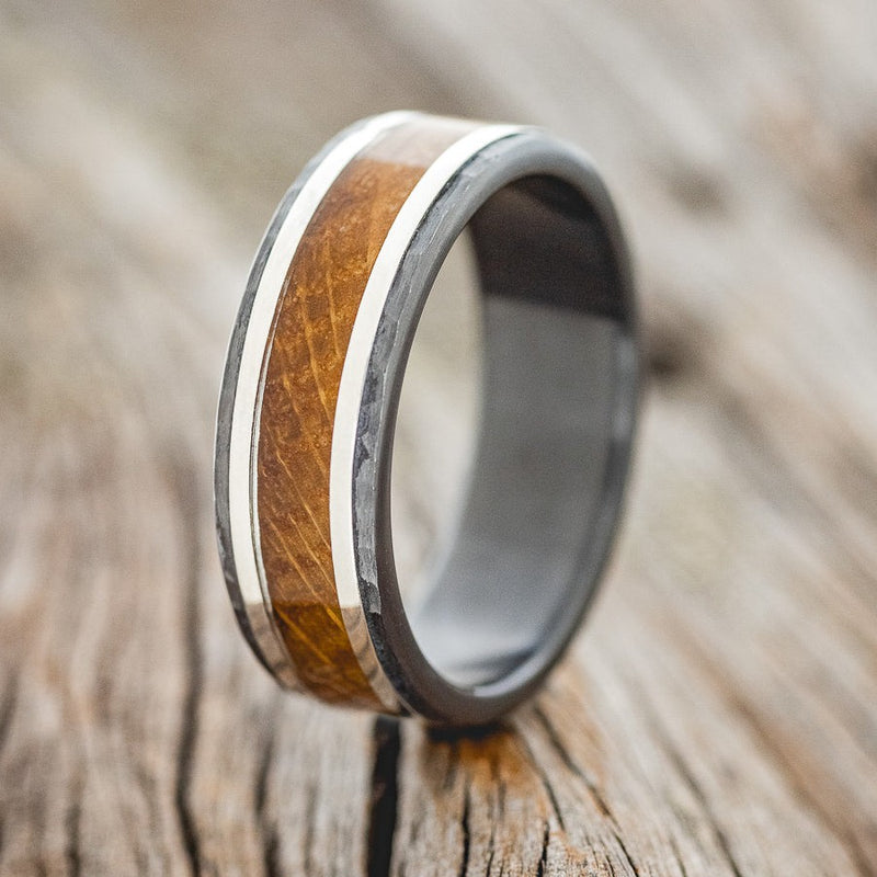 Shown here is "Hollis", a custom, handcrafted men's hammered wedding ring featuring whiskey barrel oak and silver inlays on a fire-treated black zirconium band, upright facing left. Additional inlay options are available upon request.