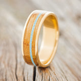 Shown here is "Rainier", a custom, handcrafted men's wedding ring featuring whiskey barrel oak and turquoise inlay on a 14K gold band, upright facing left. Additional inlay options are available upon request.