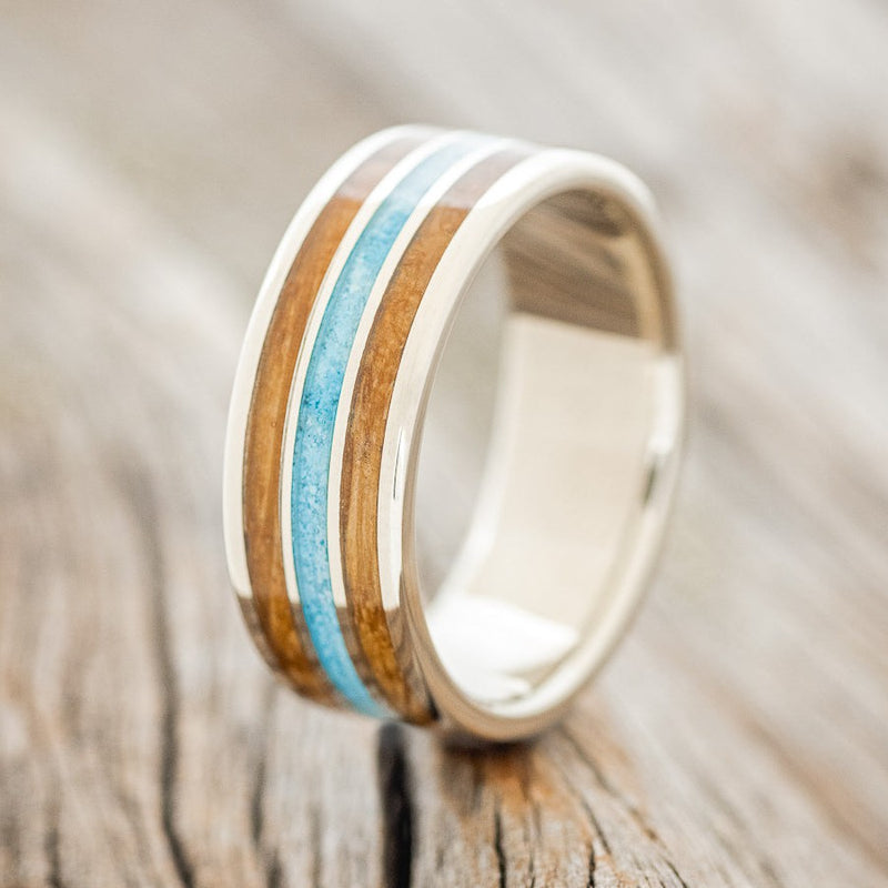 Shown here is "Rio", a custom, handcrafted men's wedding ring featuring 3 channels with a whiskey barrel and turquoise inlays on a 14K gold band, upright facing left. Additional inlay options are available upon request.