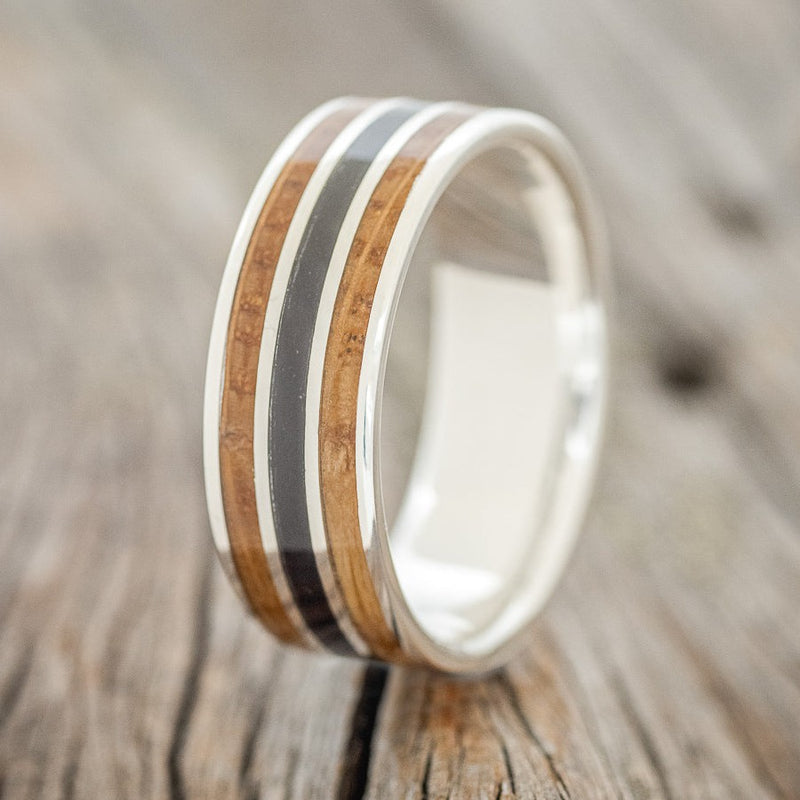 Shown here is "Rio", a custom, handcrafted men's wedding ring featuring 3 channels with a whiskey barrel and charred whiskey barrel inlay on a silver band, upright facing left. Additional inlay options are available upon request.