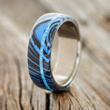 Shown here is "Remmy", a custom, handcrafted men's wedding ring featuring a blue and black G10 wave overlay and a blue opal inlay on a titanium band, upright facing left. Additional inlay options are available upon request.