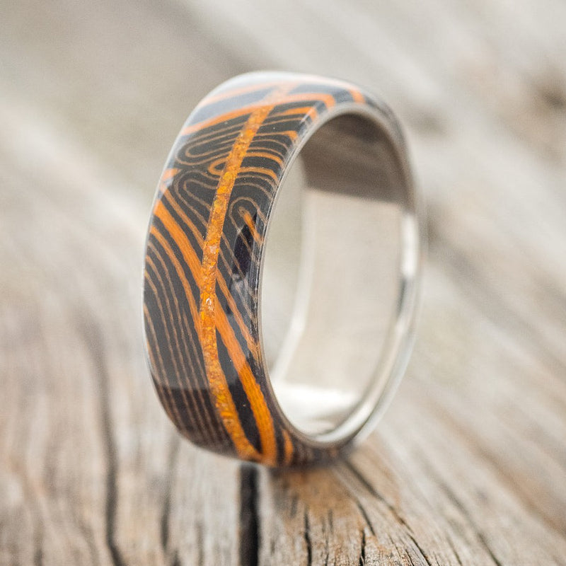 Shown here is "Remmy", a custom, handcrafted men's wedding ring featuring an orange and black G10 wave overlay and an orange opal inlay on a titanium band, upright facing left. Additional inlay options are available upon request.