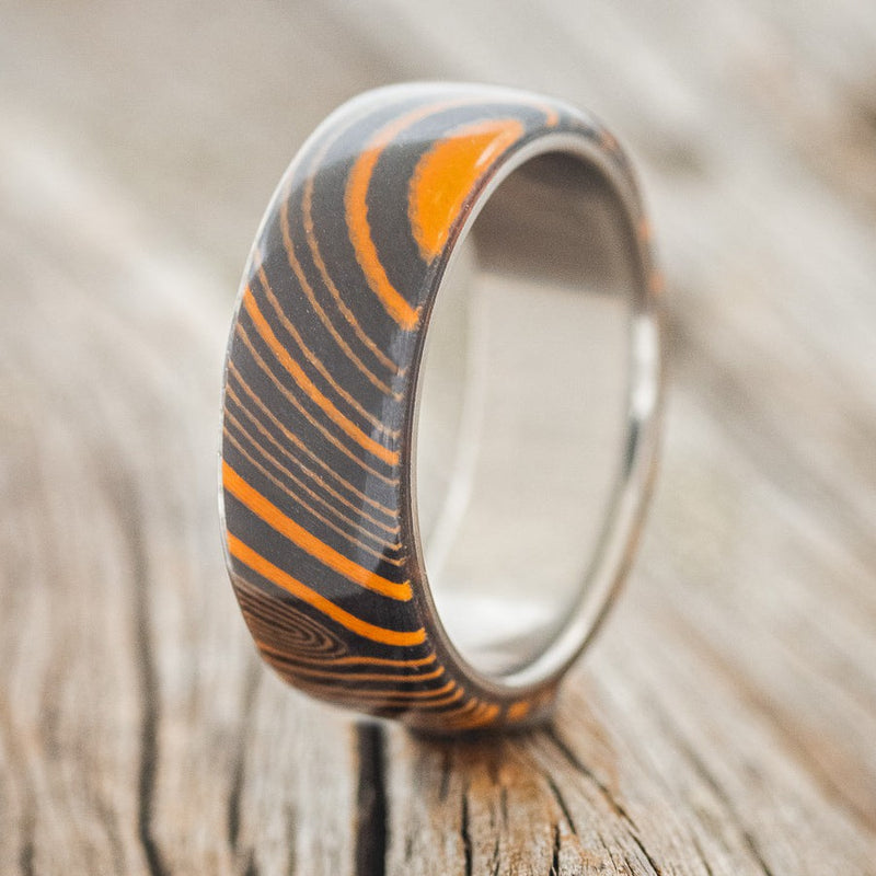 Shown here is "Haven", a custom, handcrafted men's wedding ring featuring an orange and black G10 wave overlay, upright facing left. Additional inlay options are available upon request.