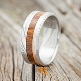 Shown here is "Tanner", a custom, handcrafted men's wedding ring featuring koa wood and a hammered 14K rose gold inlay on an etched Damascus steel band, upright facing left. Additional inlay options are available upon request.