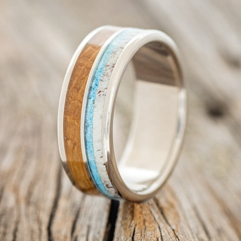 Shown here is "Dyad", a custom, handcrafted men's wedding ring featuring 2 channels with turquoise, elk antler, and whiskey barrel inlays on a titanium band, upright facing left. Additional inlay options are available upon request.