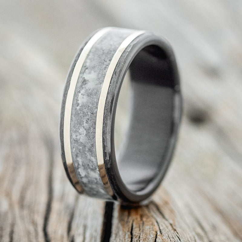 Shown here is "Hollis", a custom, handcrafted men's wedding ring featuring crushed moonstone and two 14K white gold (1mm) inlays, on a hammered, fire-treated black zirconium band, upright facing left. Additional inlay options are available upon request.